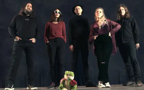 Cinq personnes se tiennent devant un fond sombre. Au premier plan, une grenouille en peluche tient une rose. Le groupe, comme un **crippled black phoenix**, semble prêt à s'envoler.