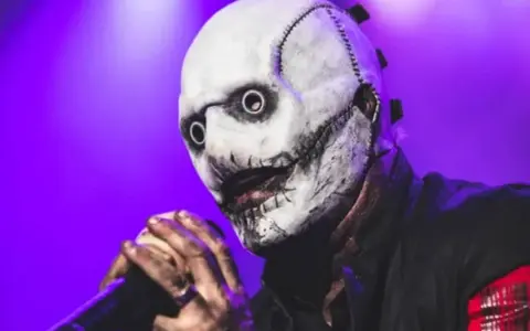 Le chanteur Slipknot, masqué et terrifiant, tient un micro sur scène sous une lumière violette.