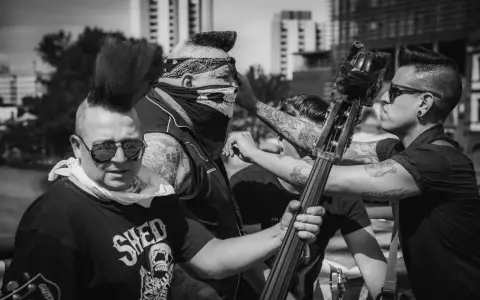 Quatre musiciens au look punk, dont un avec un bandana et un masque de crâne, se préparent à jouer. Un d'eux tient une contrebasse, prêt à faire un mad sin.