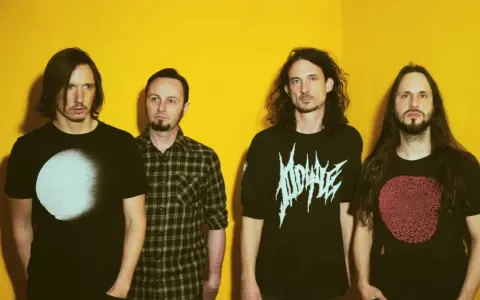 Quatre hommes aux cheveux longs posent devant un fond jaune vif. Leurs t-shirts portent des graphismes distinctifs, évoquant l'énergie brute du magma et la puissance de Gojira.