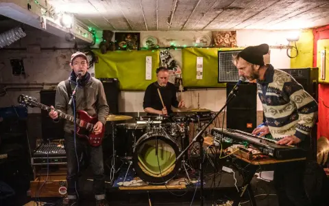 Trois musiciens jouent dans un local sombre, éclairé par des guirlandes. Un guitariste, un batteur et un claviériste, oi boys !