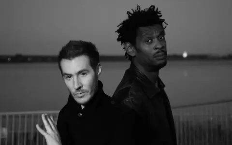 Les membres de Massive Attack, 3D et Daddy G, posent dos à dos, regardant dans des directions opposées, dans une ambiance sombre et contemplative.