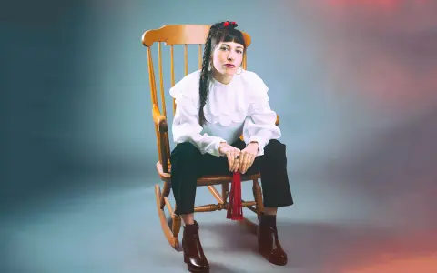 Scout Niblett, assise sur un fauteuil à bascule, porte une blouse blanche à volants et un pantalon noir. Elle tient un pompon rouge.