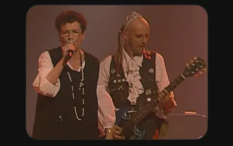 Une chanteuse et un guitariste, peut-être les ramoneurs de menhirs, sur scène. L'homme porte une couronne et une tenue excentrique.