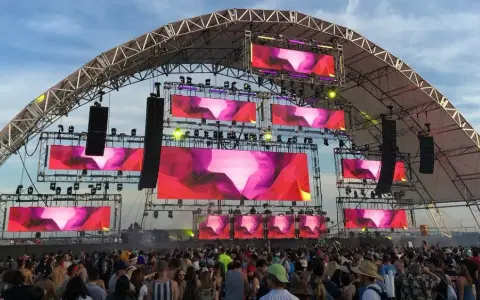 Scène de festival avec écrans LED diffusant des visuels abstraits roses et violets. Une foule danse sous la structure, attendant leur artiste deep house préféré.