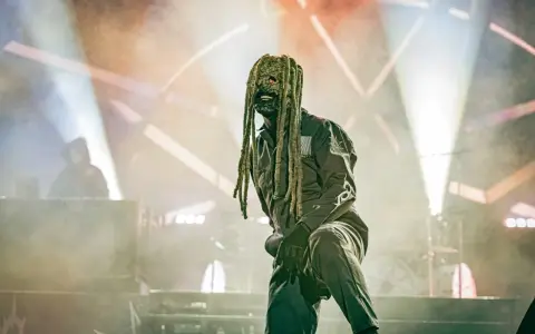 Un membre de Slipknot, masqué et en combinaison, sur scène sous des projecteurs.