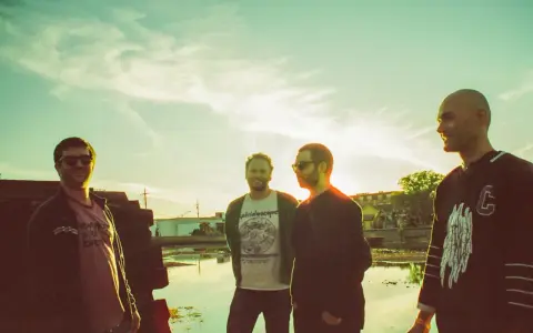 Quatre hommes posent devant un ciel clair, comme un chat pile. L'un porte un t-shirt "kaleidoscope".