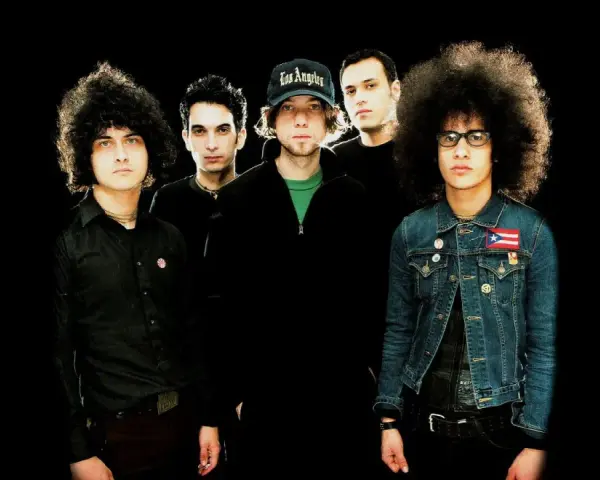 Cinq musiciens, dont certains avec des coiffures afro, posent pour une photo de groupe. L'un porte une casquette "Los Angeles". Ils sont prêts pour une soirée **at the drive in**.