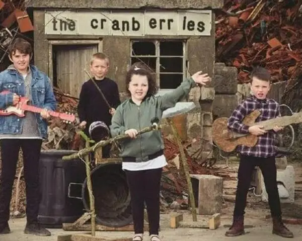 Quatre enfants jouent de la musique devant un panneau "the cranberries". L'un tient une guitare rose, un autre une guitare en bois.