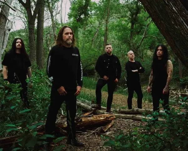 Cinq hommes en noir posent dans une forêt. Le groupe, qui pourrait être Cattle Decapitation, se tient parmi les arbres et la végétation luxuriante.