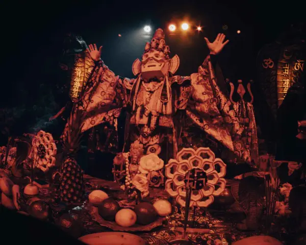 Un personnage masqué, drapé dans des tissus ornés, semble présider un rituel. Autour de lui, des offrandes de fruits, des serpents stylisés et des symboles sacrés évoquent un culte du feu mystérieux.