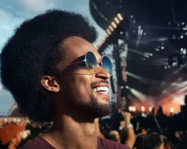 Un homme souriant avec des lunettes de soleil profite d'un concert en plein air. Il porte des bouchons d'oreilles, se demandant pourquoi pas de bouchon concert pour protéger son audition.