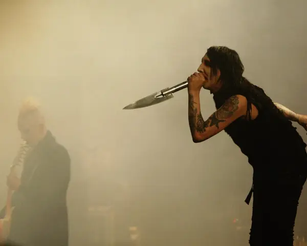 Marilyn Manson chante dans un micro en forme de couteau, un autre musicien est flou à gauche. L'ambiance d'un concert de Marilyn Manson album.