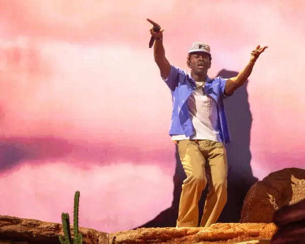 Tyler Okonma, micro en main, danse sur scène devant un décor rose pastel. Il porte une casquette, une chemise bleue ouverte sur un t-shirt blanc et un pantalon beige.