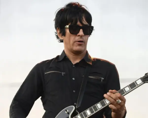 Un des membres de Eagles of Death Metal, guitariste en chemise noire et lunettes de soleil, joue sur scène.
