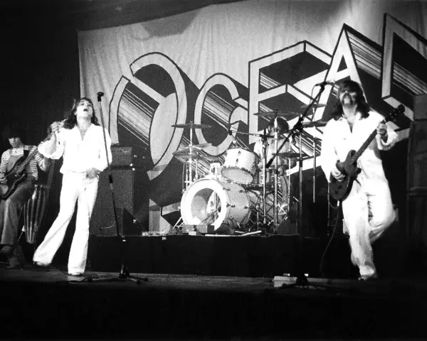 Le groupe Trust en concert, avec le chanteur en blanc au centre, un guitariste à gauche et un autre guitariste à droite. La scène est décorée d'une grande bannière avec le logo du groupe.