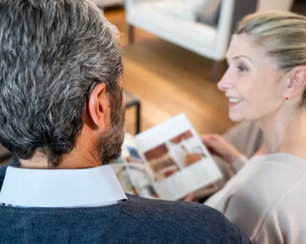 Un homme avec un appareil auditif, peut-être souffrant d'acouphènes après concert, discute avec une femme lisant un magazine.