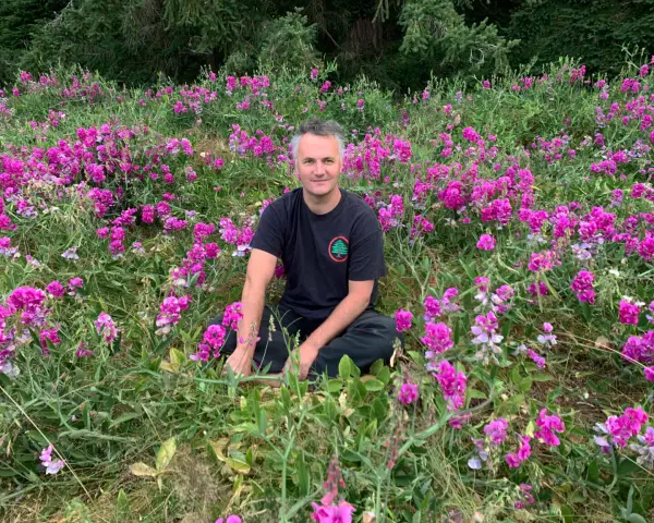 Un homme est assis au milieu d'un champ de fleurs roses et violettes, avec des arbres en arrière-plan. Il semble se trouver sur le mont Eerie.