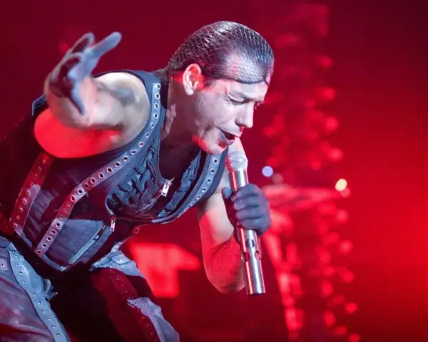 Till Lindemann, chanteur du groupe metal allemand Rammstein, en pleine performance sur scène, micro à la main, sous un éclairage rouge intense.