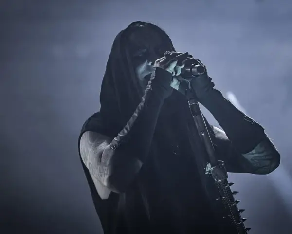Chanteur de Dimmu Borgir, visage encapuchonné et maquillé, chantant dans un micro, ambiance sombre et enfumée.