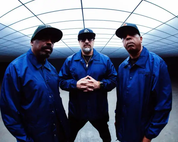 Les membres de Cypress Hill, B-Real, Sen Dog et DJ Muggs, posent ensemble, portant des vestes bleues assorties sous un plafond géométrique.