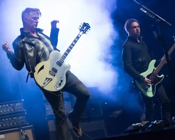 Deux membres de Queens of the Stone Age en pleine performance, l'un jouant de la guitare, l'autre de la basse, sous des éclairages bleus intenses.
