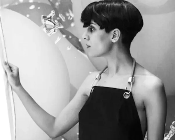 Fatima Al Qadiri, artiste à la coupe courte, porte un tablier noir, regardant une œuvre abstraite.