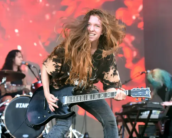 Reba Meyers, guitariste au look sauvage, fait vibrer sa guitare électrique sur scène, cheveux au vent, devant un fond rougeoyant.
