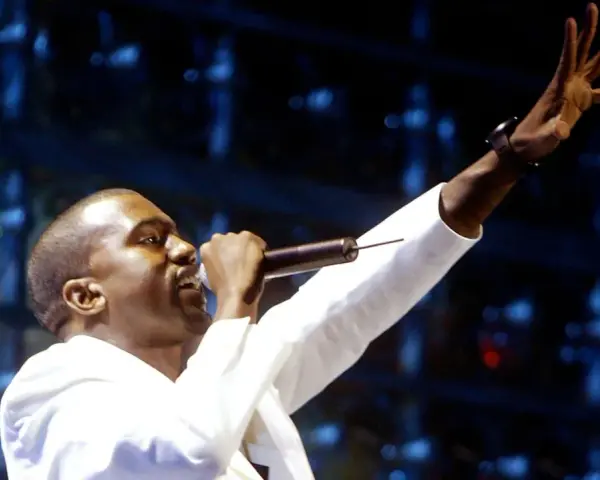 Kanye West, aux origines de sa carrière, chante sur scène, micro à la main, bras levé.