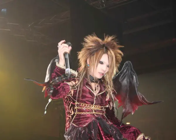 Un membre de X Japan, cheveux ébouriffés, maquillage prononcé, costume sombre et ailes de démon, chante sur scène.