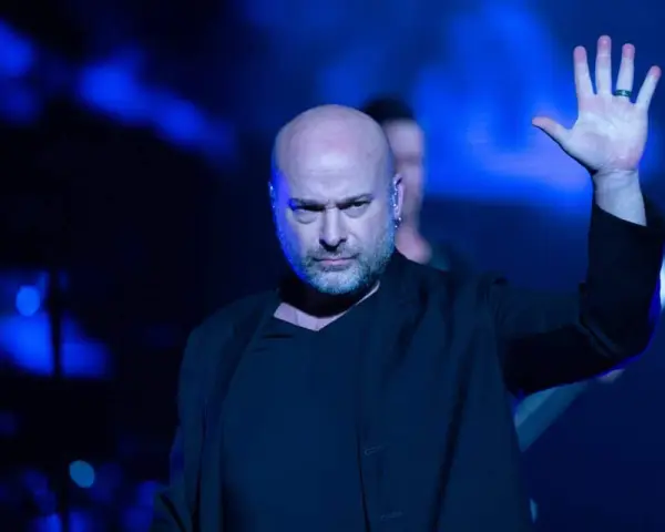 Le chanteur Disturbed, chauve et barbu, lève la main sur scène sous des lumières bleues intenses.