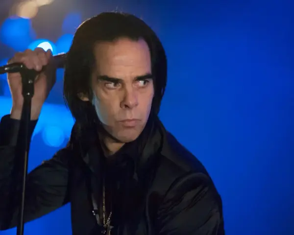 Nick Cave, chanteur du groupe australien des années 80, sur scène, micro à la main, regard intense sur fond bleu.