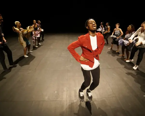 Un danseur afro 93, en veste rouge, mène une troupe sur scène. Les danseurs, aux tenues variées, exécutent des mouvements synchronisés.