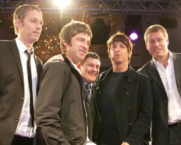Cinq hommes, dont le groupe Oasis, posent ensemble sur scène, souriants sous les lumières.