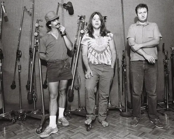 Trois hommes dans un studio d'enregistrement, entourés de micros. L'un d'eux, casquette et short, se touche la moustache. Husker du, un souvenir de cette époque.