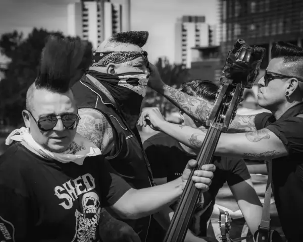 Quatre musiciens au look punk, dont un avec un bandana et un masque de crâne, se préparent à jouer. Un d'eux tient une contrebasse, prêt à faire un mad sin.