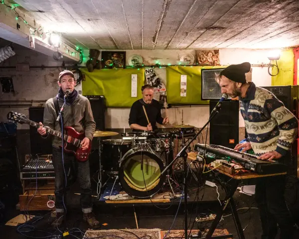 Trois musiciens jouent dans un local sombre, éclairé par des guirlandes. Un guitariste, un batteur et un claviériste, oi boys !