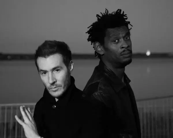 Les membres de Massive Attack, 3D et Daddy G, posent dos à dos, regardant dans des directions opposées, dans une ambiance sombre et contemplative.