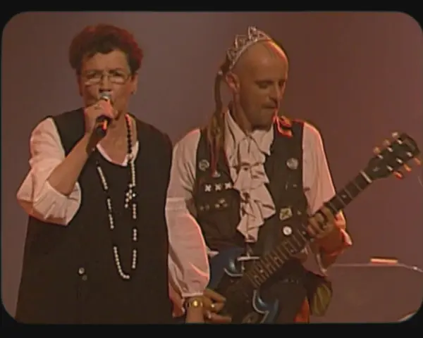 Une chanteuse et un guitariste, peut-être les ramoneurs de menhirs, sur scène. L'homme porte une couronne et une tenue excentrique.