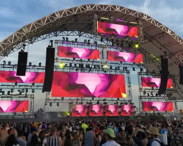 Scène de festival avec écrans LED diffusant des visuels abstraits roses et violets. Une foule danse sous la structure, attendant leur artiste deep house préféré.