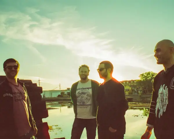 Quatre hommes posent devant un ciel clair, comme un chat pile. L'un porte un t-shirt "kaleidoscope".