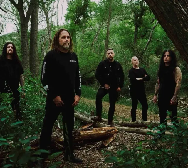 Cinq hommes en noir posent dans une forêt. Le groupe, qui pourrait être Cattle Decapitation, se tient parmi les arbres et la végétation luxuriante.
