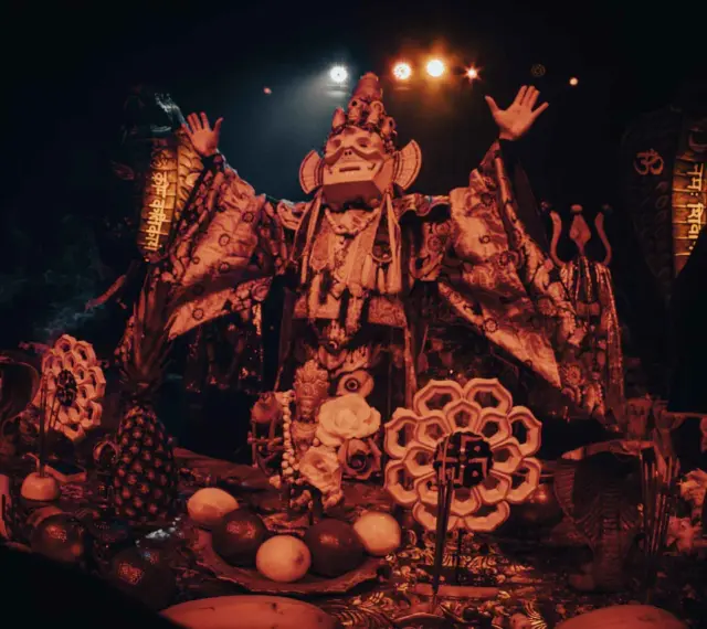 Un personnage masqué, drapé dans des tissus ornés, semble présider un rituel. Autour de lui, des offrandes de fruits, des serpents stylisés et des symboles sacrés évoquent un culte du feu mystérieux.