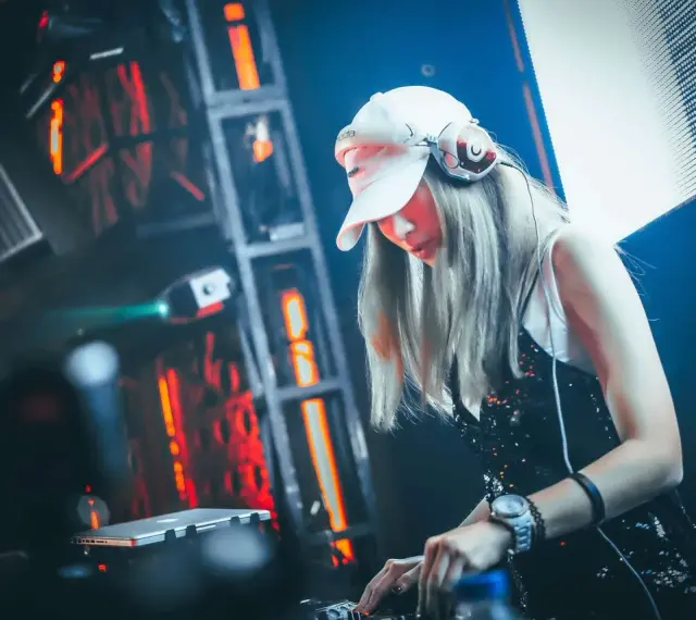 Une dj femme, casquette blanche et casque sur les oreilles, mixe dans une ambiance lumineuse et électrique.