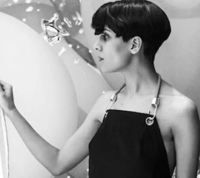Fatima Al Qadiri, artiste à la coupe courte, porte un tablier noir, regardant une œuvre abstraite.