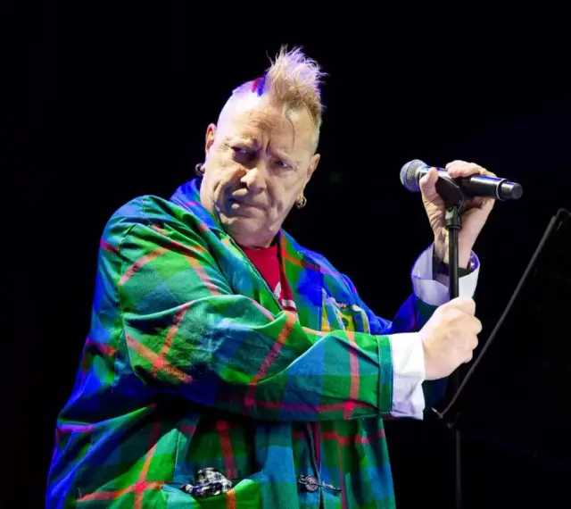 Le chanteur des Sex Pistols, John Lydon, avec sa crête iconique, porte une veste à carreaux écossais colorée et tient un microphone.