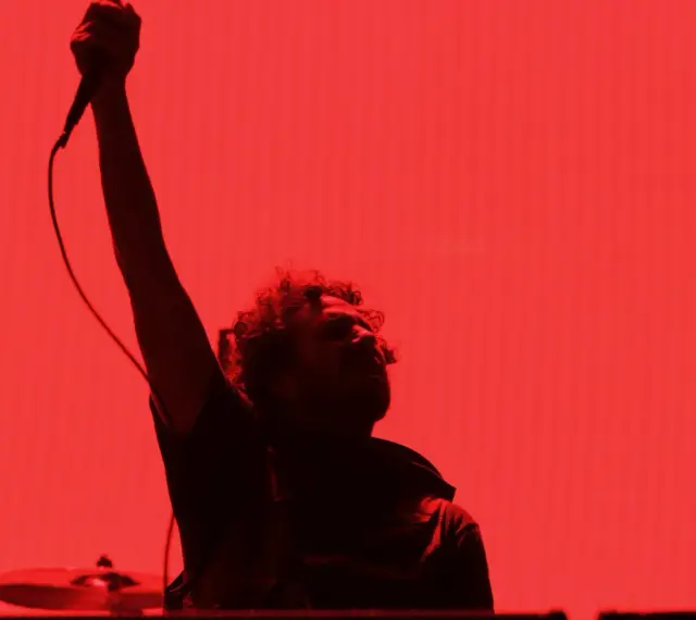 Chanteur de Rage Against the Machine, micro levé, sur fond rouge vif. L'énergie de leurs albums en live.
