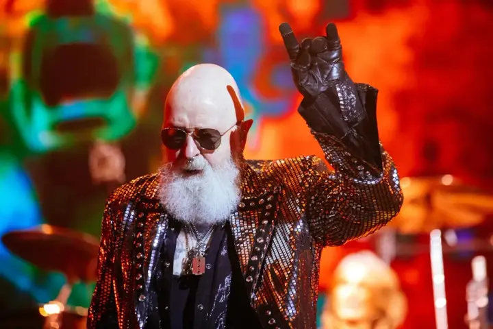Rob Halford, un des membres de Judas Priest, fait le signe des cornes sur scène, vêtu d'une veste scintillante.