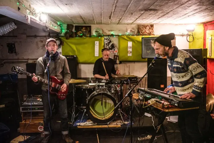 Un groupe de musique punk des années 90 joue dans un local sombre. Un guitariste, un batteur et un claviériste sont sur scène.