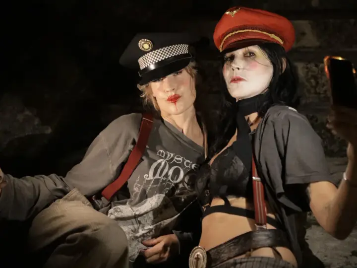 Deux femmes aux maquillages théâtraux portent des casquettes et des bretelles, l'une tenant un téléphone.
